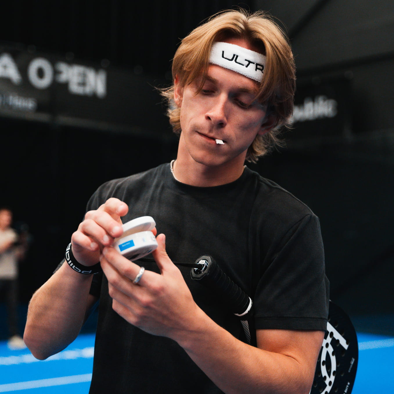 A man on a padel court holding a white round container with a small white pouch in his mouth.